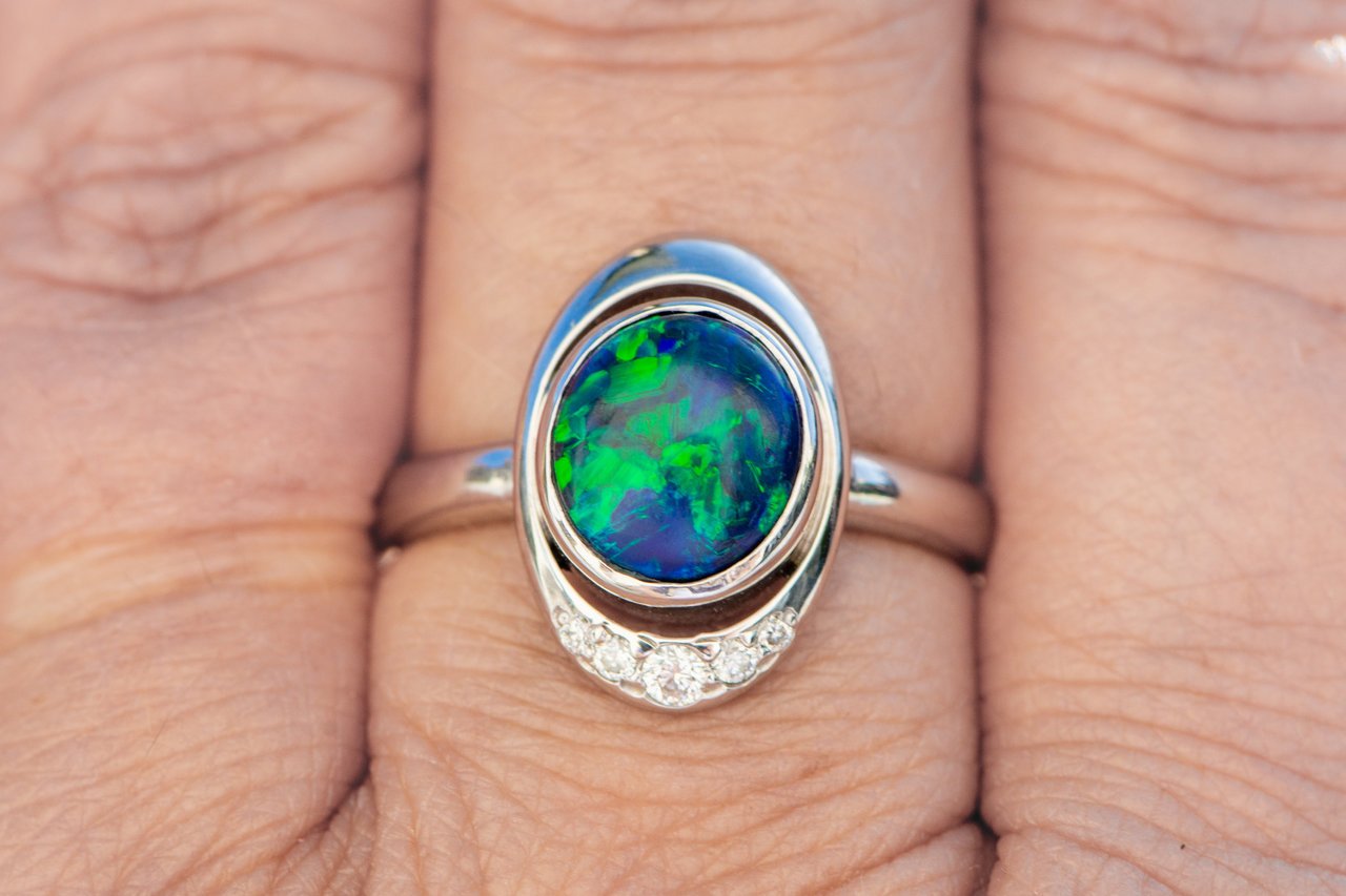 Punks-Tricks posted this marvelous black opal ring on the Show Me the Bling forum at PriceScope. The stone was a birthday present and her goal was to capture the essence of the aurora borealis, and add a peekaboo emerald to honor her Mother. I’d say she massively succeeded, because this is phenomenal!