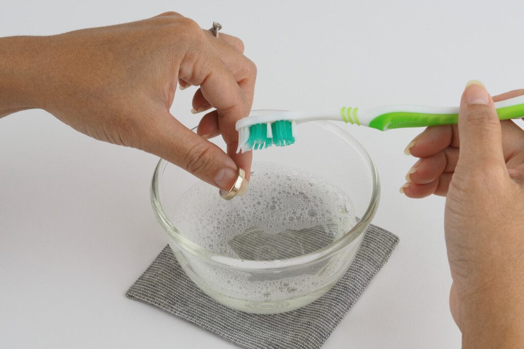 A silver ring being cleaned using a toothbrush and soapy water.