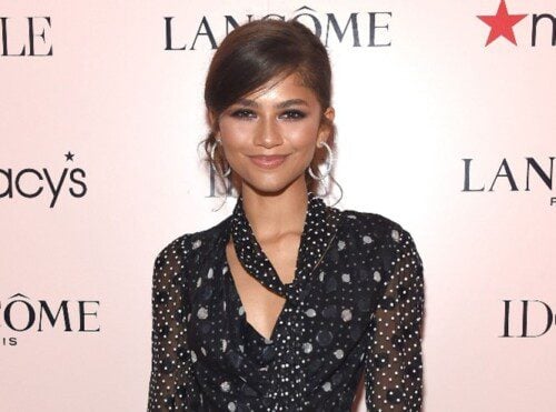 Zendaya facing forward, in black with white polka dots, loose scarf that matches blouse, side swept bangs, in front of Lancome Red Carpet Background. Diaomnd Hoop Earrings.