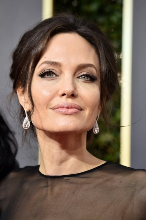 Close up of Angelina Jolie, slightly smiling. Sheer black top, hair up, diamond teardrop earrings, nude lip, heavy eye makeup