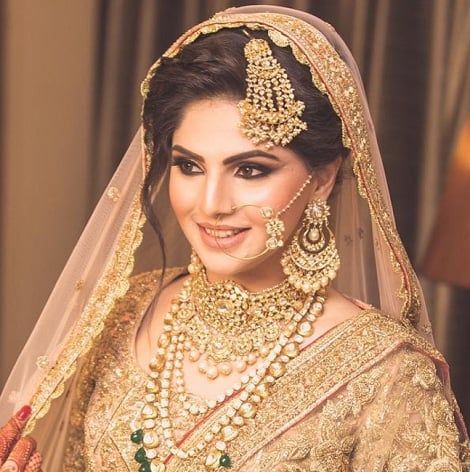 Bride in golden veil with gold jewelry, smiling. Maang Tikka Passa, Nosering, earrings, stacked necklaces