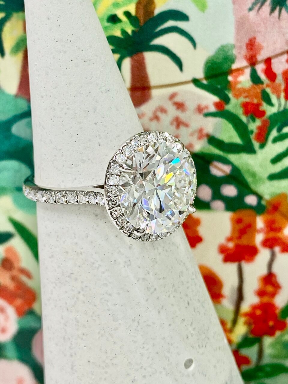 On a ring stand with a brightly floral backdrop, a custom cut 5ct Whiteflash ACA diamond ring is displayed.