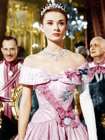 Audrey Hepburn in a ballroom, from the film Roman Holiday. She is in a pink off the shoulder gown with a darker pink sash, a tiara, diamond necklace, and multiple diamond brooches. She has one full length white opera glove.