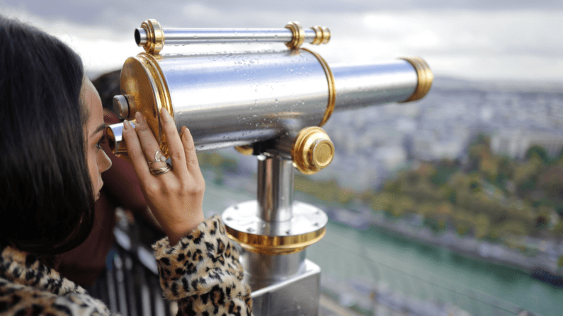 Young woman of indeterminate race in leopard print looks into a telescope that is faced at a city in the distance. She is wearing rings that are visible.