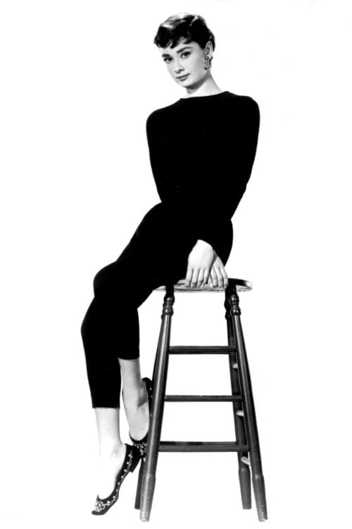 Picture of a woman in all black, black hair in chignon, cigarette pants, sitting on a stool. Wearing ballet flats. Audrey Hepburn