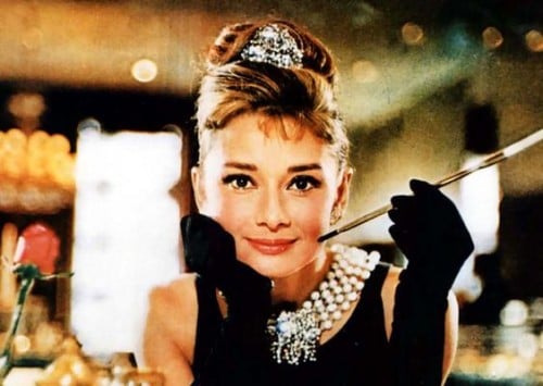 The classic image from Breakfast at Tiffany's. Store faded behind her while she has an updo, a tiara, a black dress, a large pearl and diamond necklace, black opera gloves. She had her face leaned into her hand and she is smoking a cigarette with a long cigarette holder.