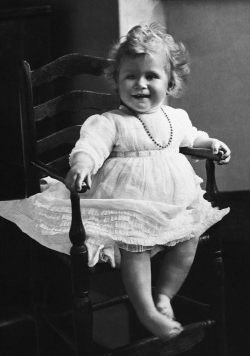 Greyscale image of 1 year old Queen Elizabeth II in a chair and a white dress with a necklace. She has blonde curls.