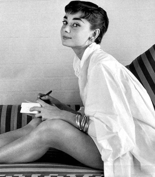 Audrey Hepburn sitting sideways on a striped sofa, image is greyscale, bare legged, pearl earrings, oversized white button down shirt, she is writing a note and has her head turned to the camera with a bit of a side eyed mischievous look.