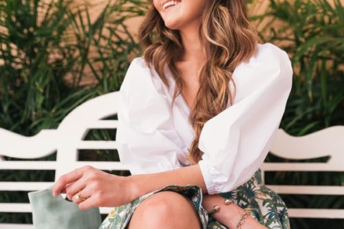 woman neck and shoulders, beach wavy hair on white button down shirt, white slate bench and trees in background. Wearing rings and bracelets.