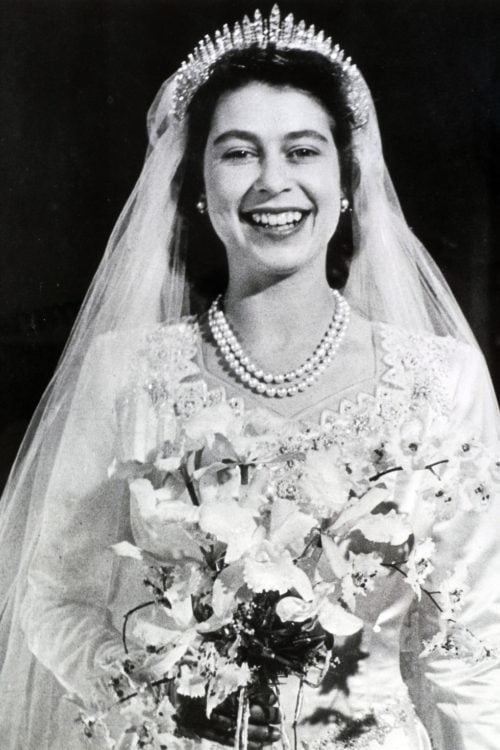 Queen Elizabeth II in her wedding gown with a bouquet of flowers a double strand of pearls and the Queen Mary Fringe Tiara with a bridal veil.