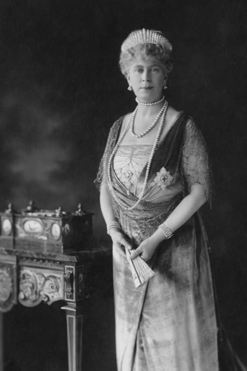 Greyscale image of Queen Mary of England standing regally decked out in a fine gown, jewels, and The Queen Mary Fringe Tiara.