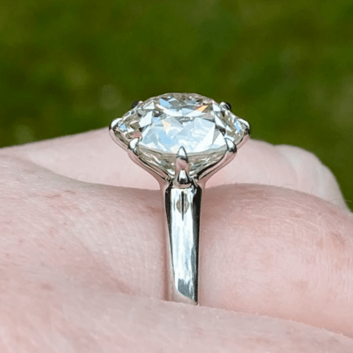 Large diamond covered in pastel light reflections on a silver toned band on a finger (though no more of the hand is visible. There is grass in the background.