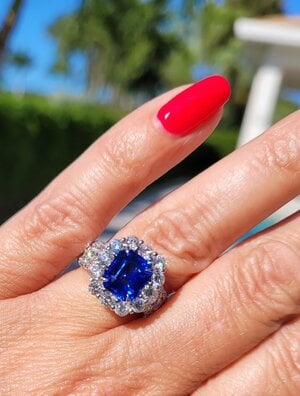 Sapphire ring with a diamond halo, long fingers with long red nails. There is a blue sky and greenery behind the hand.