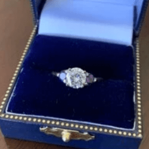 A large white center stone with two smaller purple stones in a dark blue ring box with a gold clasp for the box. On a wooden table top. 