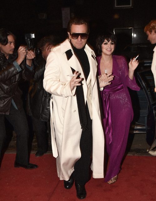 a man in a white duster over a black suit leading a brunette woman in a purple dress on a red carpet with paparazzi 