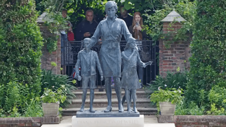 A statue of a with two children, in a gated garden, lush plants and onlookers looking over the gate in the background.