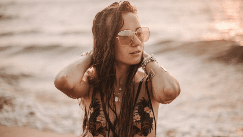 woman in surf at beach, in swimwear, jewelry, and sunglasses with her hands on either side of her head. Sort of sepia toned.