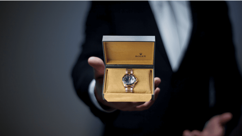 head and shoulders of a person in a tuxedo on a grey background extending a box with a rolex watch in it