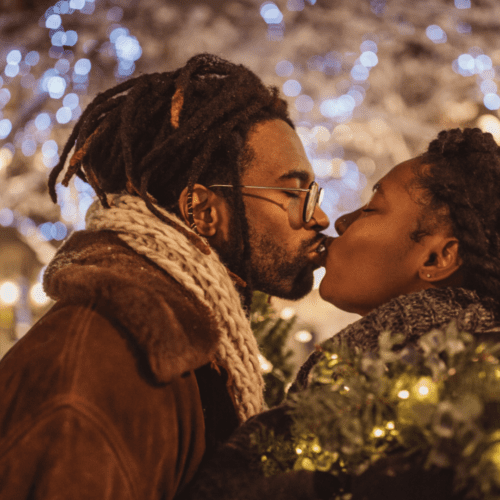 A black couple kissing among greenery and holiday lights.