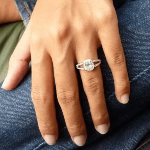 A hand against denim jeans, there is a split shank diamond ring on the ring finger.