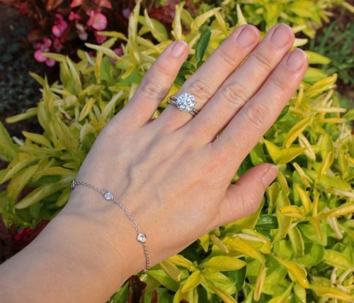 A hand over greenery, a bracelet formed with spaced out diamonds set along a chain is on the wrist, and a large diamond ring on the ting finger.