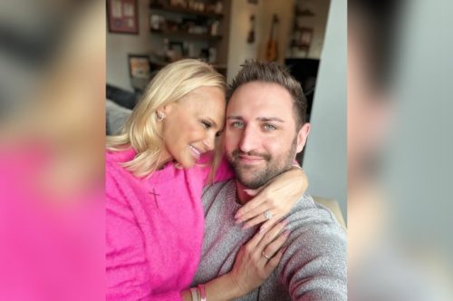 Blonde woman in pink dress her forehead against the temple of a young bearded male in grey facing the viewer.