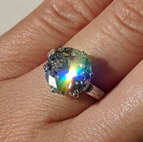 A hand with a large diamond ring with a rainbow splash of light on the head of the stone.