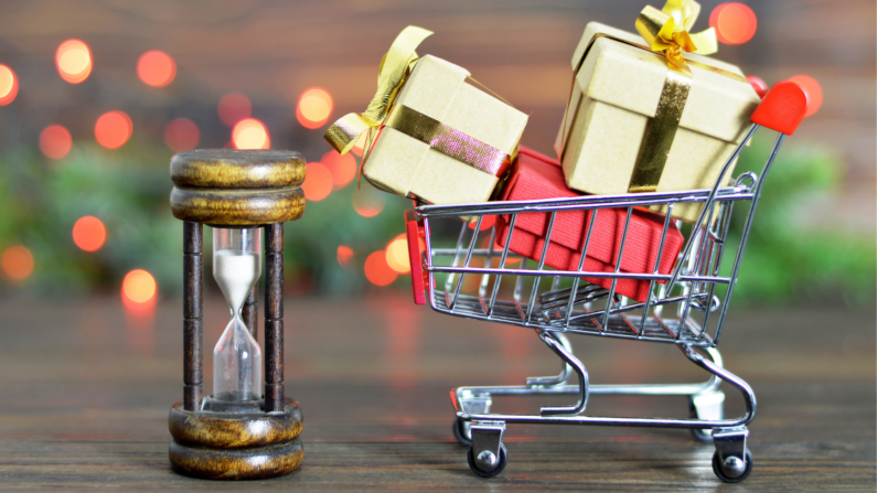 A small shopping cart full of gift boxes and an hour glass, muted holiday lights in the background.