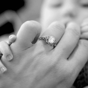 Black and white image of a hand with a diamond ring, blurred image of a baby behind and tiny little fingers wrapped around the side of the hand.