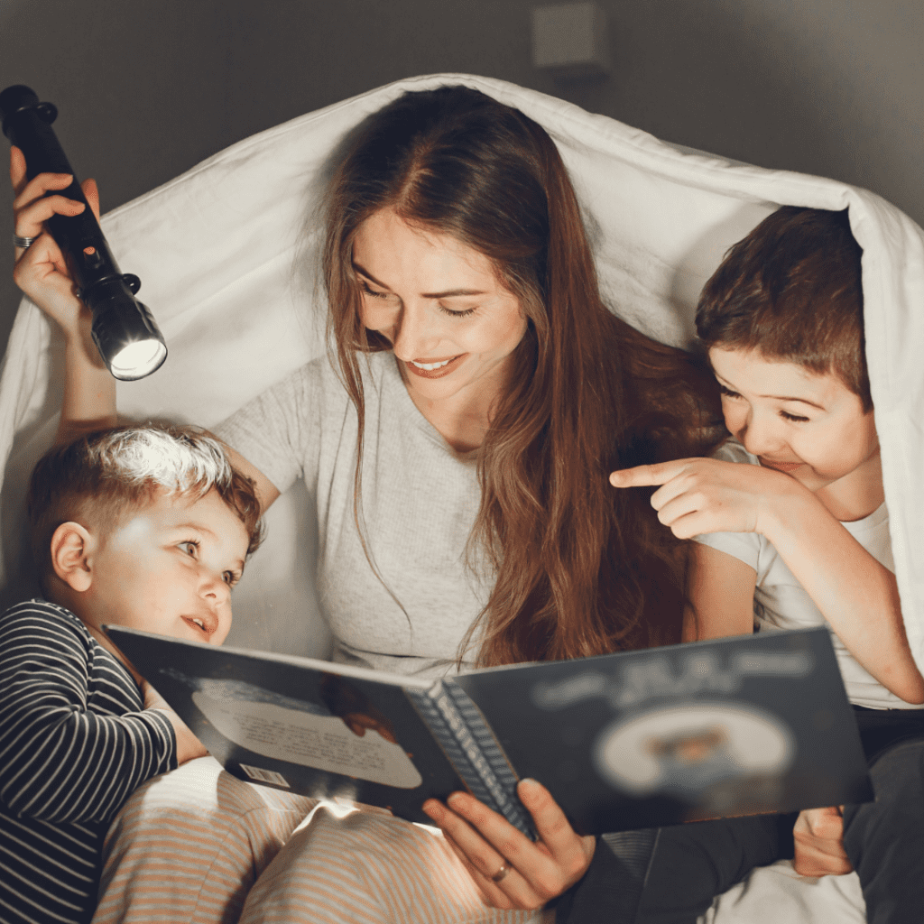 Mother and 2 small children reading under a blanket with a flash light.