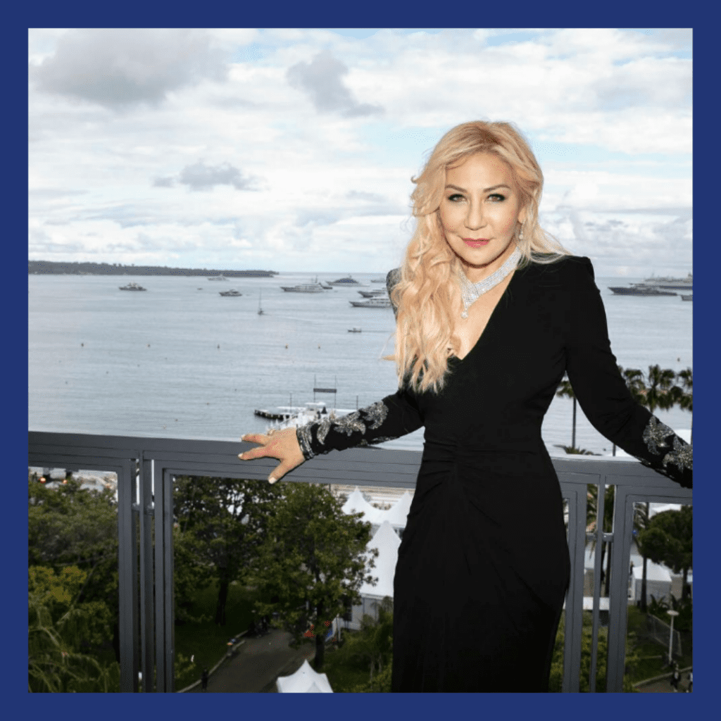 blonde woman with the sea behind her, she is wearing a lot of jewelry.