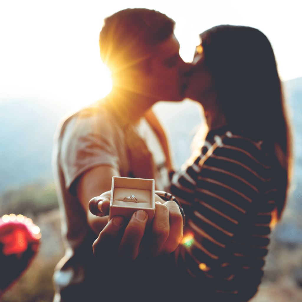 hand held out showing engagement ring with couple blurred in background