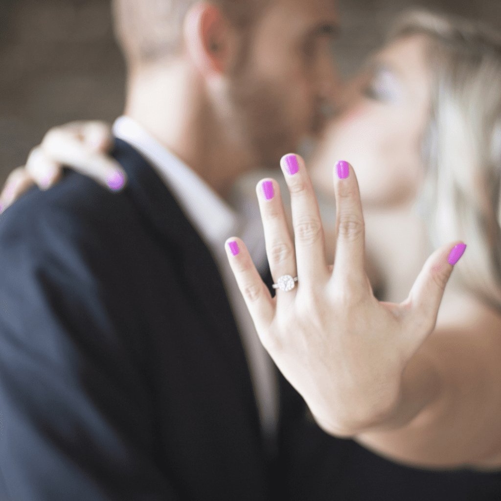 hand held out showing engagement ring with couple blurred in background