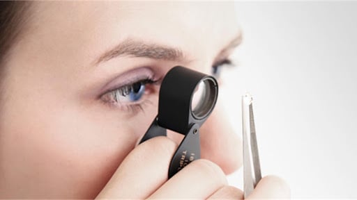 Close up image of expert examining and grading a diamond in a lab using a jeweller's loupe and tweezers