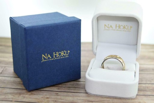 Two gift boxes, one white, and one blue, with gold foiled lettering displaying Na Hoku. Inside the white gift box, is a perfectly presented gold ring
