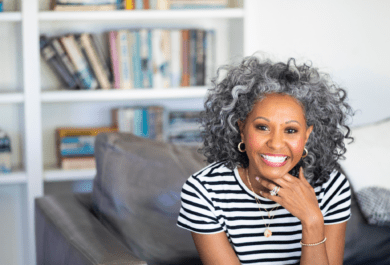 smiling woman with brown skin and salt and pepper curls sitting on a sofa in front od a bookcase. SHe is wearing hoop earrings, a couple of necklaces, and a diamond ring