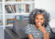 smiling woman with brown skin and salt and pepper curls sitting on a sofa in front od a bookcase. SHe is wearing hoop earrings, a couple of necklaces, and a diamond ring