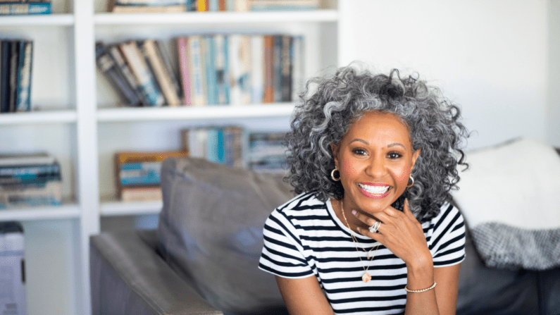 smiling woman with brown skin and salt and pepper curls sitting on a sofa in front od a bookcase. SHe is wearing hoop earrings, a couple of necklaces, and a diamond ring